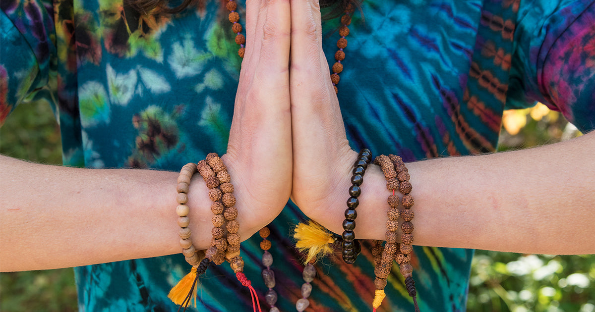 Two hands in a meditative mudra covered in mala bead bracelets with a tie dye shirt in the background