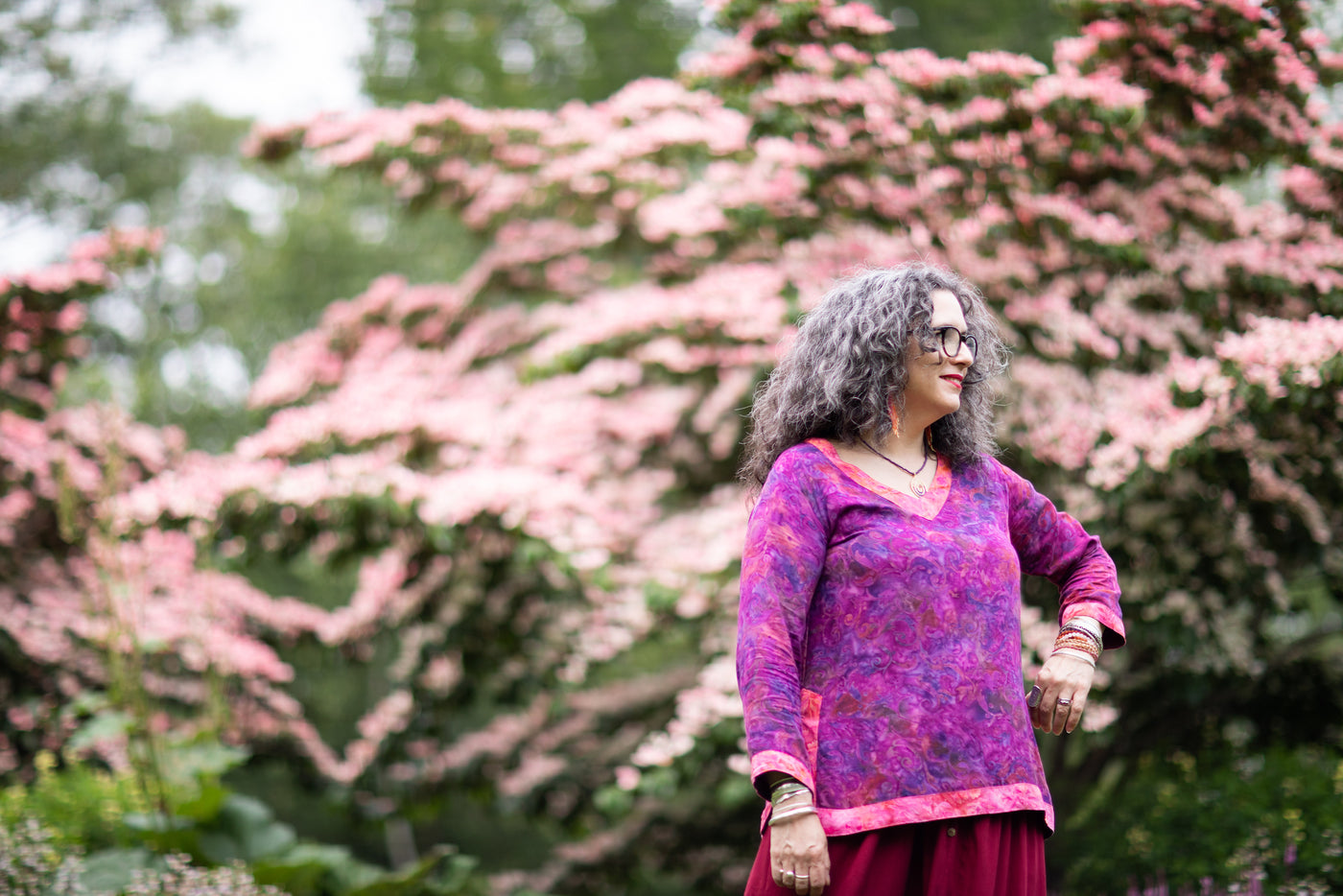 beautiful curly haired woman with graying hair and glasses wearing a bright purple two tone batik tunic in front of a flowering pink dogwood tree