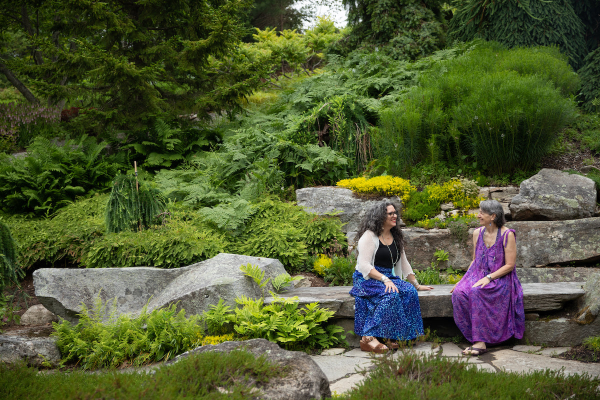 Two beautiful hippie women sitting on a stone bench in the forest in colorful Indonesian batik outfits and hippie jewelry