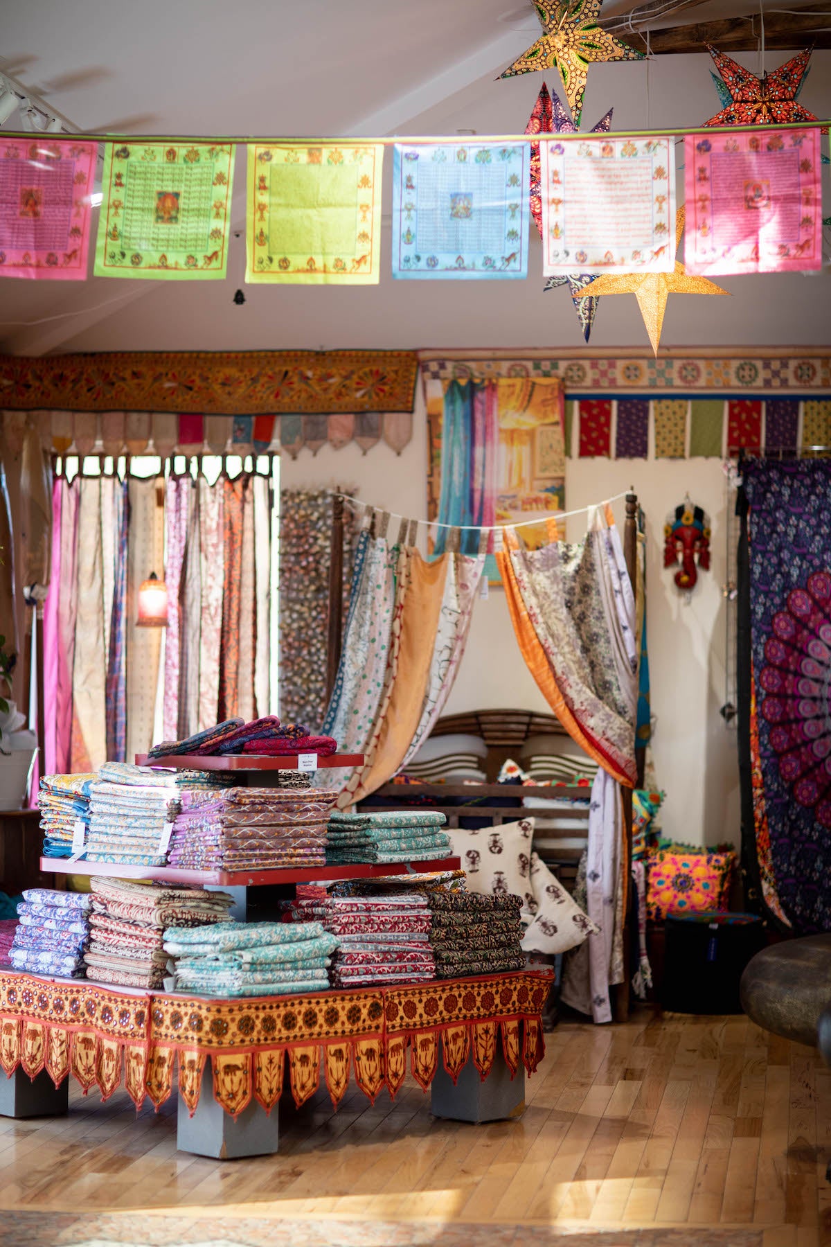 inside the beautiful hippie store that is our flagship Mexicali Blues store in Newcastle, Maine. Pictured are a pile of hand block printed tapestries, recycled silk panel curtains, prayer flags and colorful handmade hippie textiles from all over the world