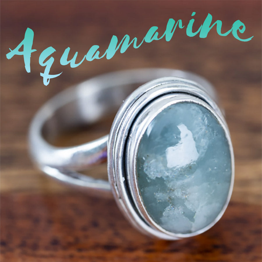 An aquamarine gemstone set in a sterling silver ring with the word "aquamarine" above it on a wood background