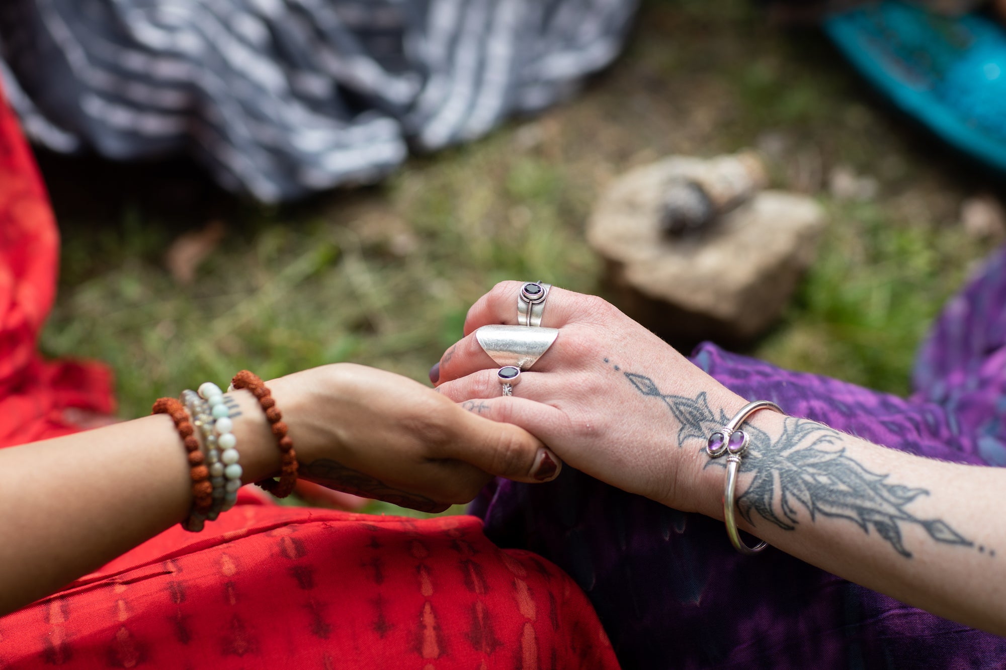 two women holding hands and meditating with arm tattoos covered in magical gemstone jewelry over a colorful backdrop