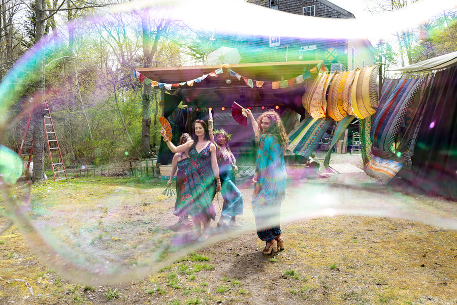A photo taken through a bubble of four beautiful hippie women with long hair dancing in front of a colorful backdrop of silk and prayer flags in colorful hippie outfits from Thailand and waving fans in the air
