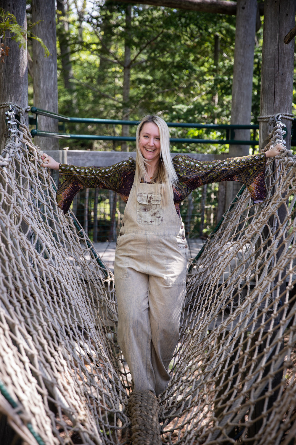 A beautiful blonde woman crossing a rope bridge in mushroom print overalls