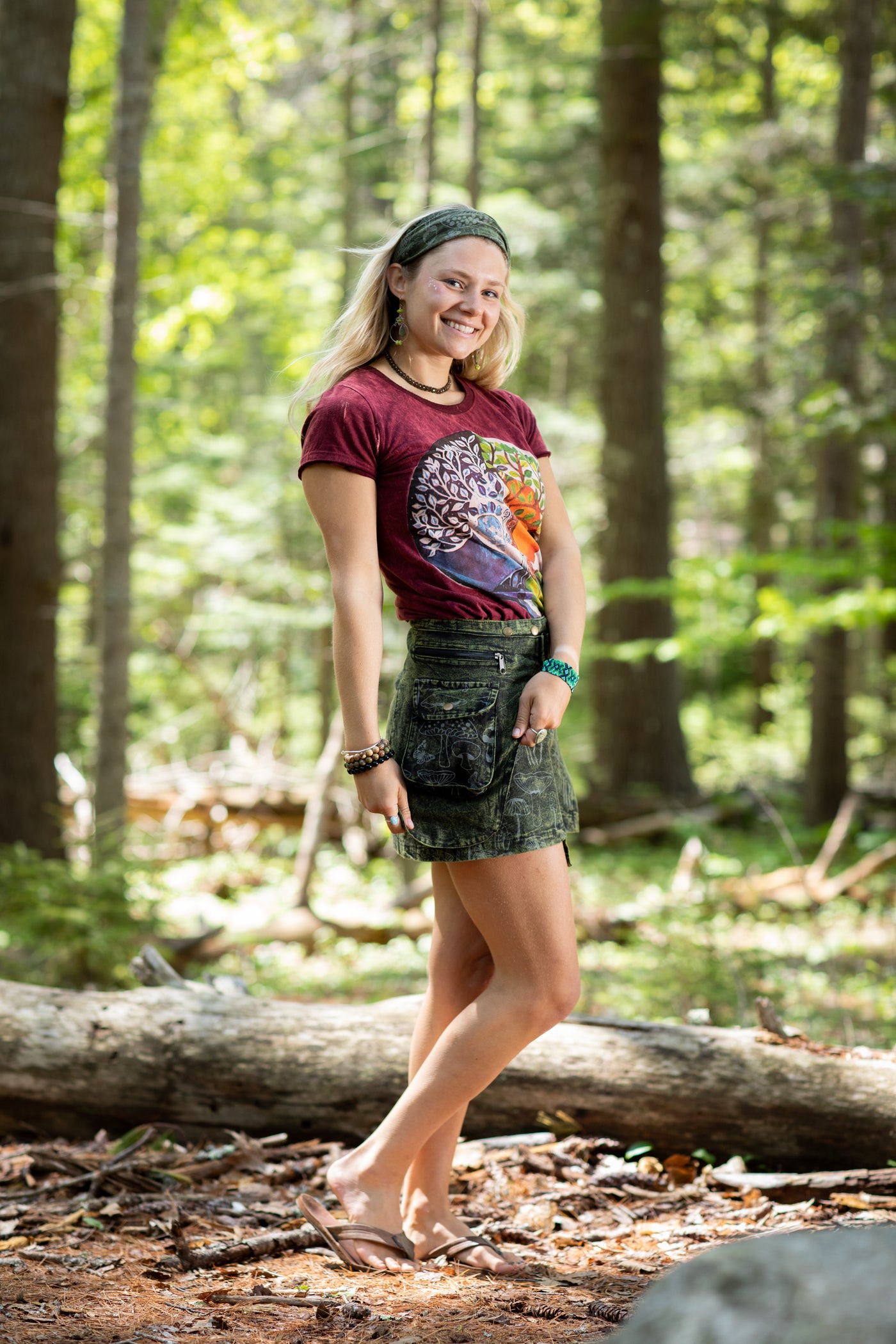 A beautiful blonde woman in the forest wearing a green mushroom headband, a maroon tree of life t shirt, and a green mushroom festival skirt