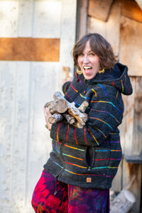 A woman outdoors wearing a dark charcoal hooded jacket with vibrant rainbow-colored horizontal stripes and holding a small bundle of firewood. She is smiling broadly, with rustic wooden and white paneled walls in the background.
