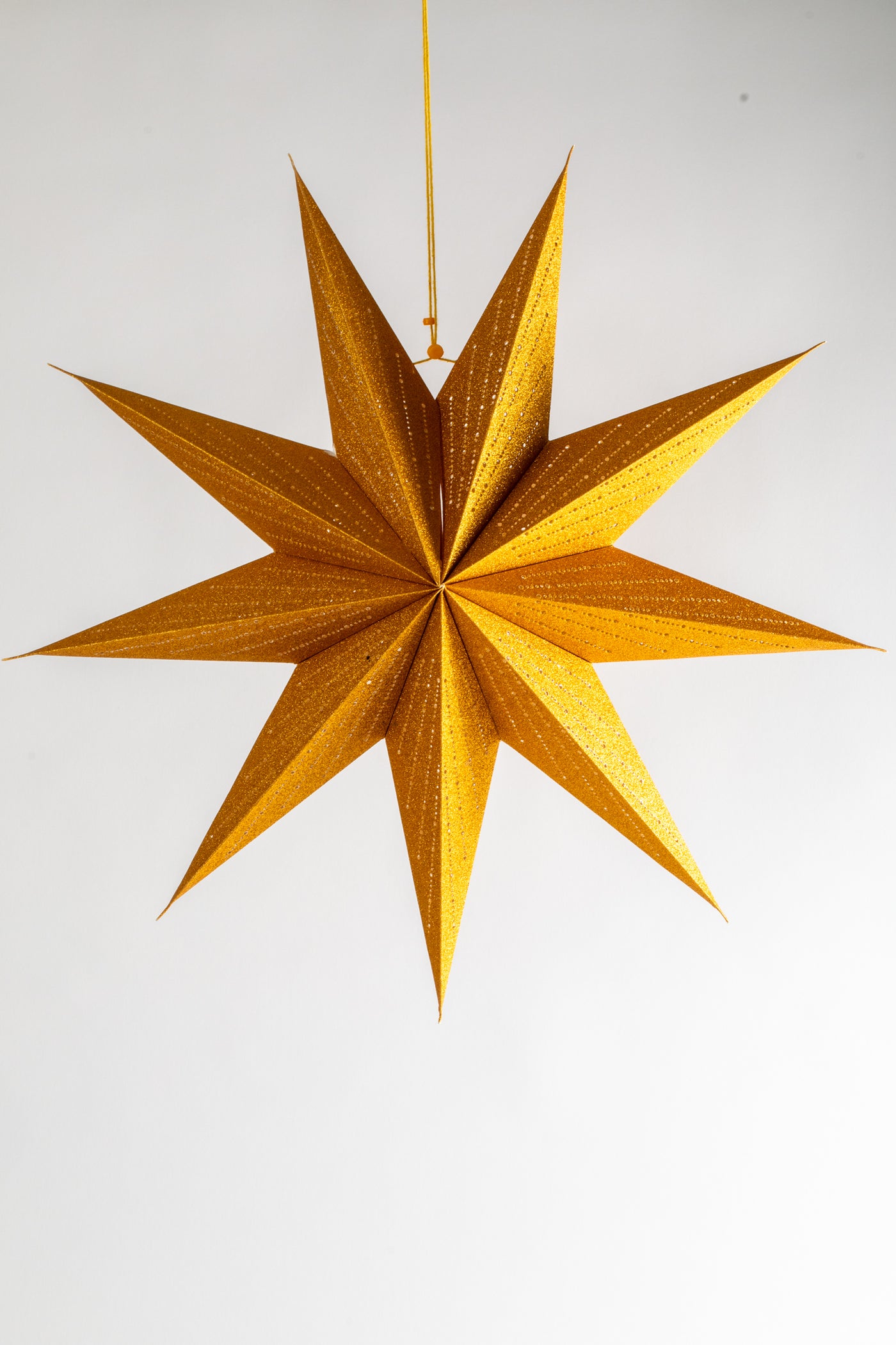 A 9-point paper star lantern in gold hanging in front of a white backdrop. 