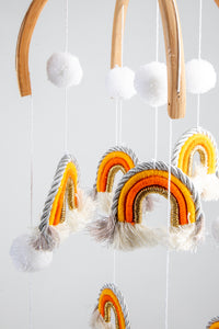 A woven rainbow mobile in Orange with orange and white rainbows and white pom-pom details, suspended from a wooden hoop with silver and gold detailing.