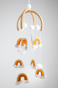 A woven rainbow mobile in Orange with orange and white rainbows and white pom-pom details, suspended from a wooden hoop with silver and gold detailing.