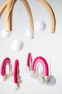A woven rainbow mobile in Pink with pink and white rainbows and white pom-pom details, suspended from a wooden hoop with silver detailing.