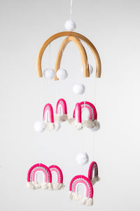 A woven rainbow mobile in Pink with pink and white rainbows and white pom-pom details, suspended from a wooden hoop with silver detailing.