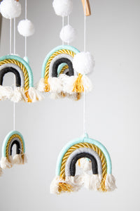 A woven rainbow mobile in Teal with blue and white rainbows and white pom-pom details, suspended from a wooden hoop with gold detailing.