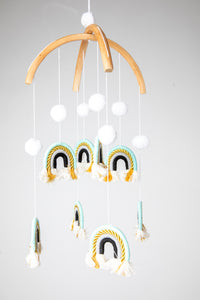 A woven rainbow mobile in Teal with blue and white rainbows and white pom-pom details, suspended from a wooden hoop with gold detailing.