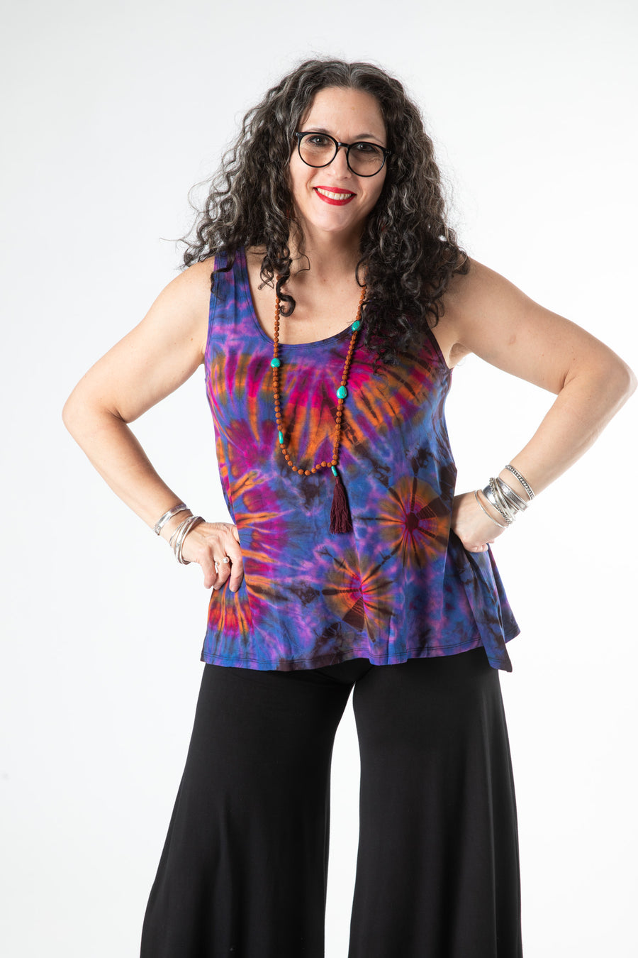 Smiling boho woman wearing a colorful purple tie dye tank top with black wide leg pants and a long hippie necklace 