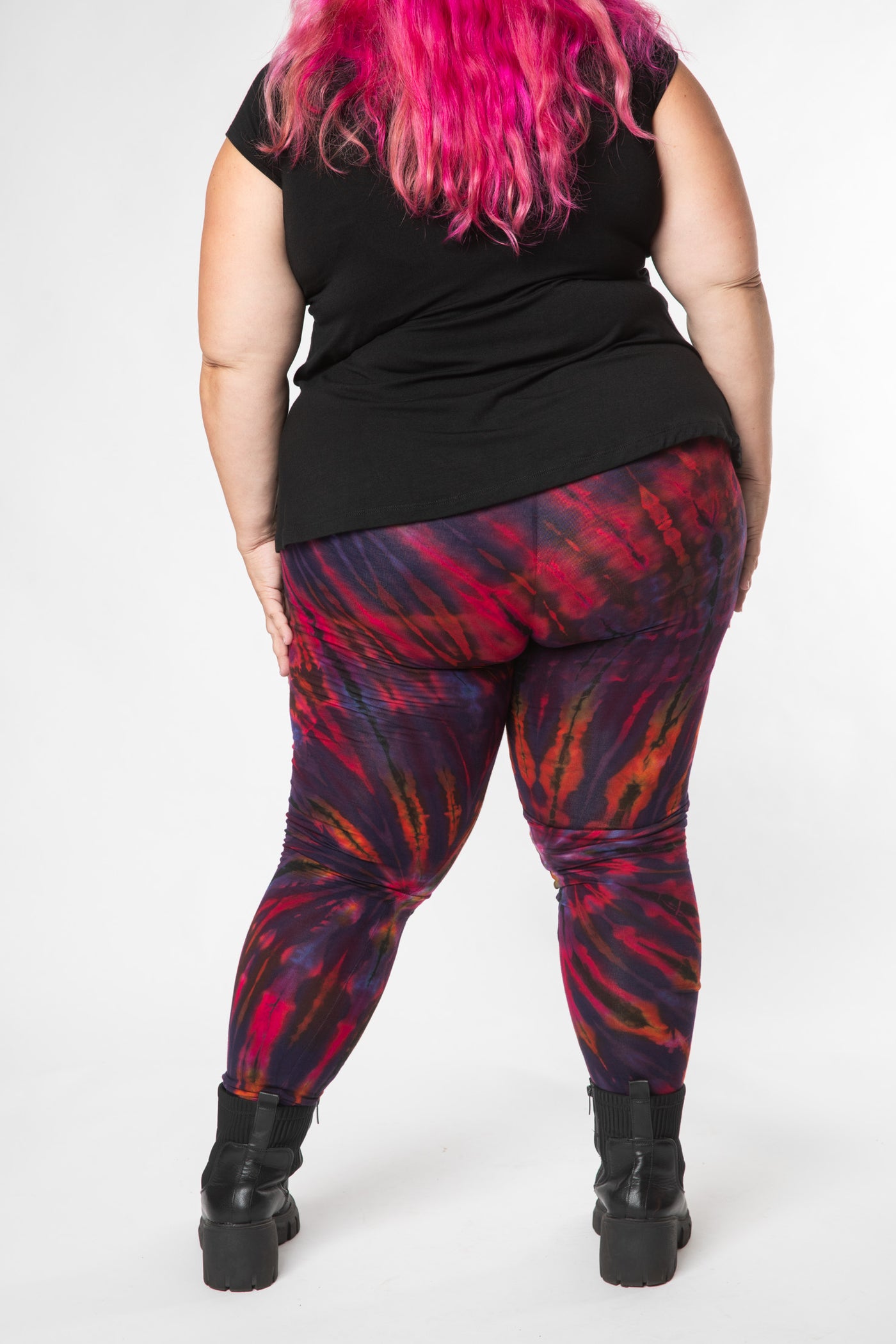 Hippie woman with pink hair facing backwards, wearing colorful tie dye leggings in purple with a black top and black boots 