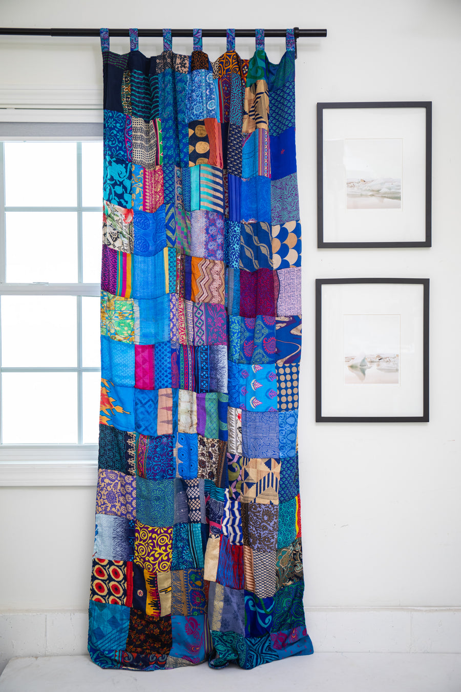Colorful patchwork curtain in Blue hanging in front of a window with framed pictures on the wall.
