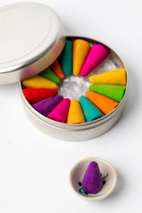 Purple incense cone displayed in ceramic holder beside open tin of rainbow incense cones