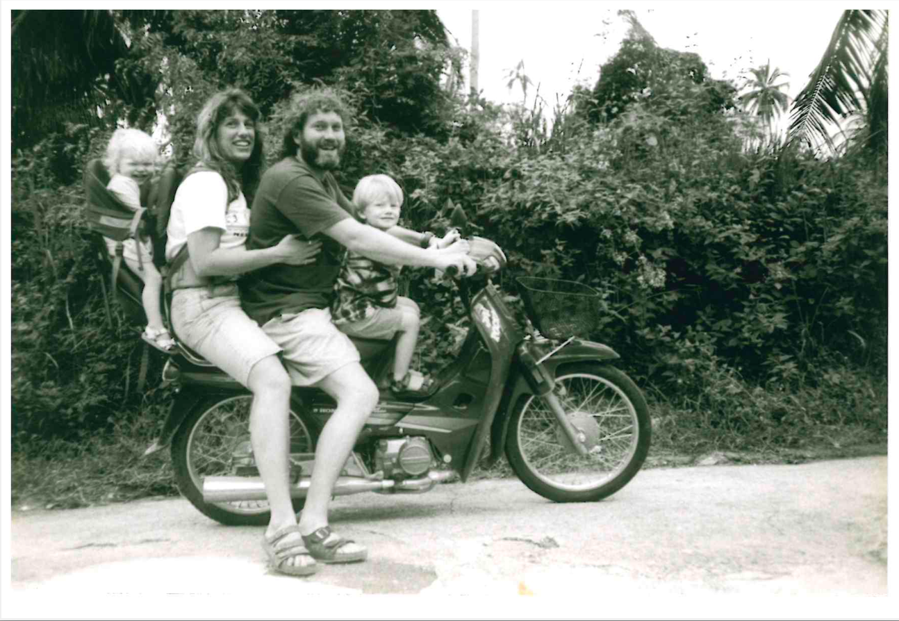 A black and white photo of family of four all on one motorbike in Thailand, a blonde infant in a baby carrier, a beautiful mother holding onto her husband and a blonde child in the front, all wearing sandals with long hair