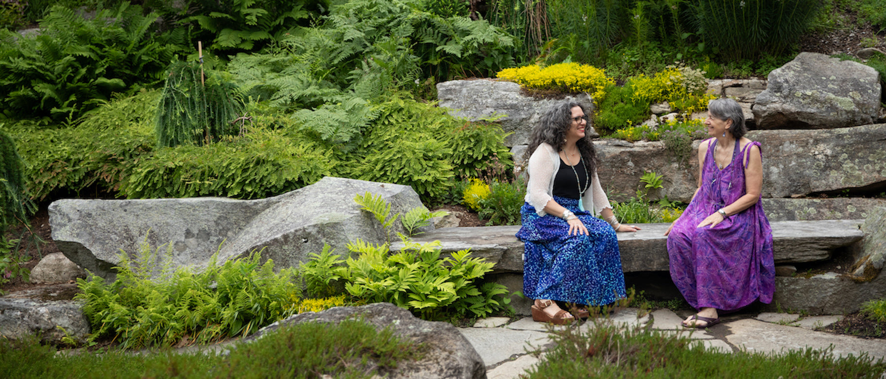 Two beautiful hippie women sitting on a stone bench in the forest in colorful Indonesian batik outfits and hippie jewelry