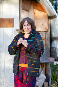 A woman wearing a dark charcoal zip-up jacket with vibrant multicolor stripes in red, yellow, green, and blue, layered over a patterned top and colorful pants, standing outdoors in front of a rustic wooden shed.