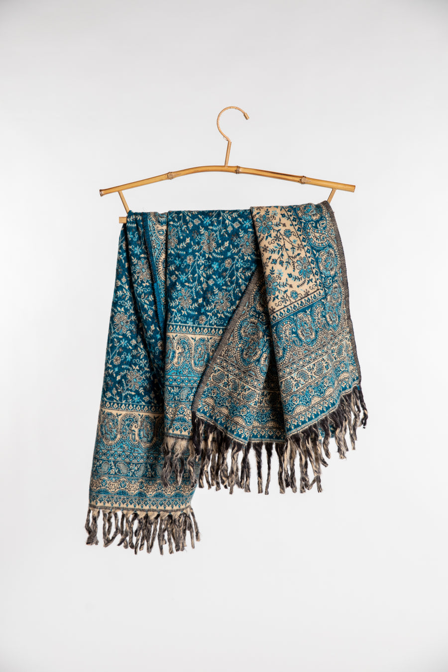 A blue paisley soft brushed shawl with fringe, draped over a bamboo hanger against a light gray background.