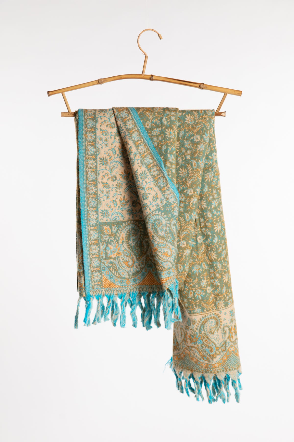 The green paisley soft brushed shawl draped over a bamboo hanger against a clean white background. The scarf features a teal and beige paisley design with turquoise fringe.