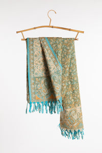 The green paisley soft brushed shawl draped over a bamboo hanger against a clean white background. The scarf features a teal and beige paisley design with turquoise fringe.