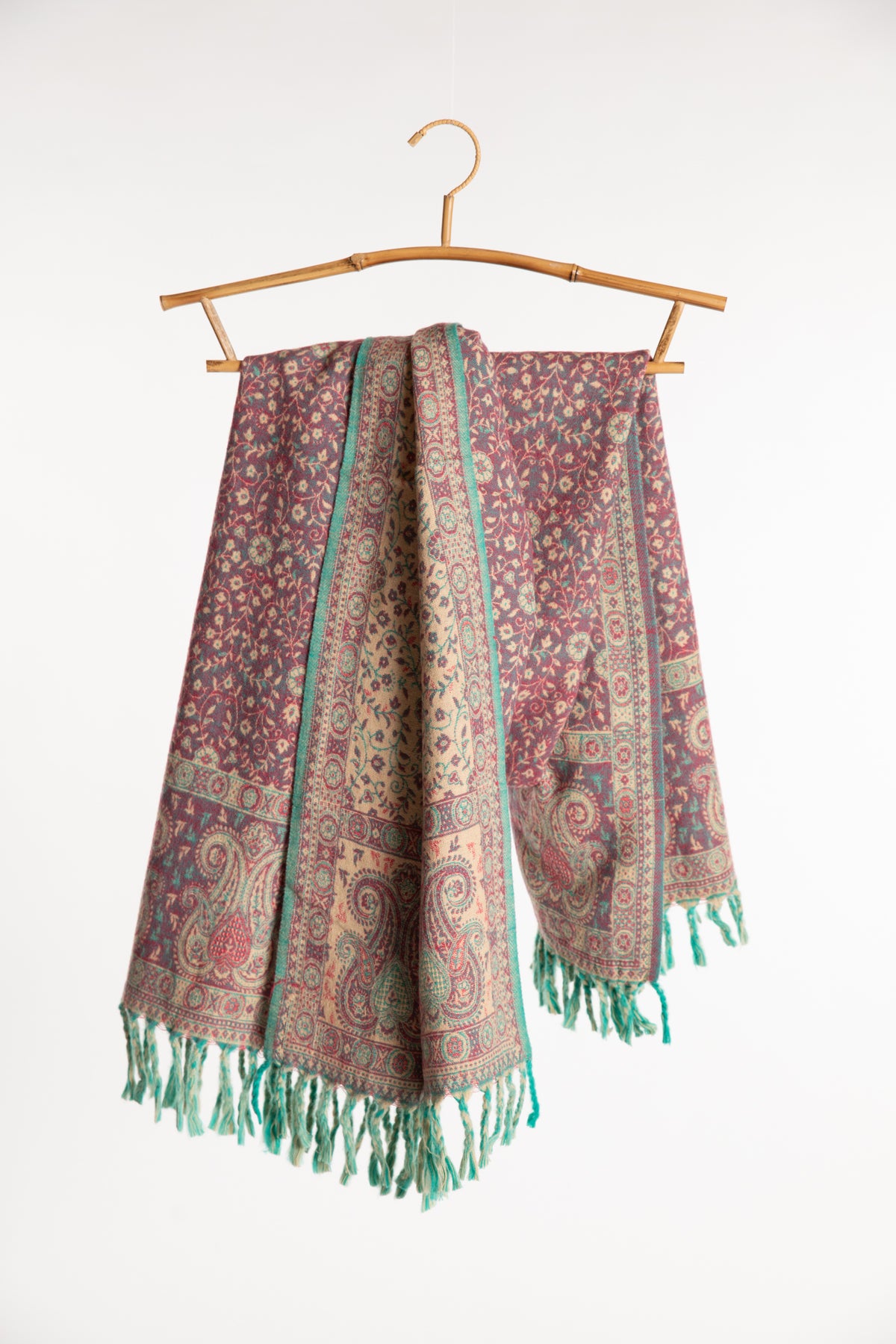 The rose paisley soft brushed shawl with green fringe, draped over a bamboo hanger against a plain white background.