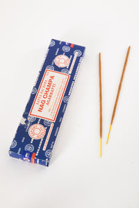 A box of Satya Sai Baba Nag Champa incense with two incense sticks placed in front of it on a white background.