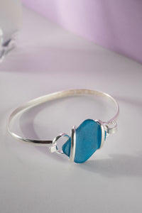 A wrapped alpaca sea glass bracelet with blue sea glass and silver-colored metal wrapping, displayed against a light purple background.