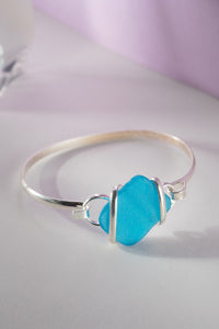 A wrapped alpaca sea glass bracelet with blue sea glass and silver-colored metal wrapping, displayed against a light purple background.